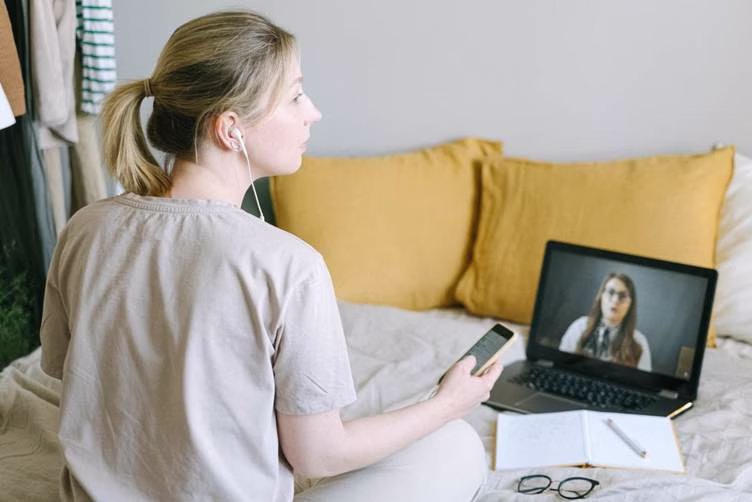 image - Alaska Sleep Clinic - Alaska Sleep Clinic Person wearing earbuds sits on a bed, talking to someone on a laptop during a video call with a notebook and glasses nearby.